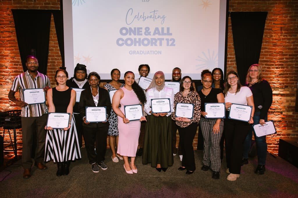 LEAP's One & All Cohort 12 graduates pose on a stage holding their certificates showing completion of the program.