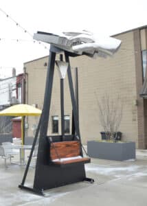 A picture of a sculpture called Keeping Time located in TIme Piece Park, Charlotte Michigan shows a metal hand playing a keyboard sitting on top of a bench that moves the keyboard when sitting. 
