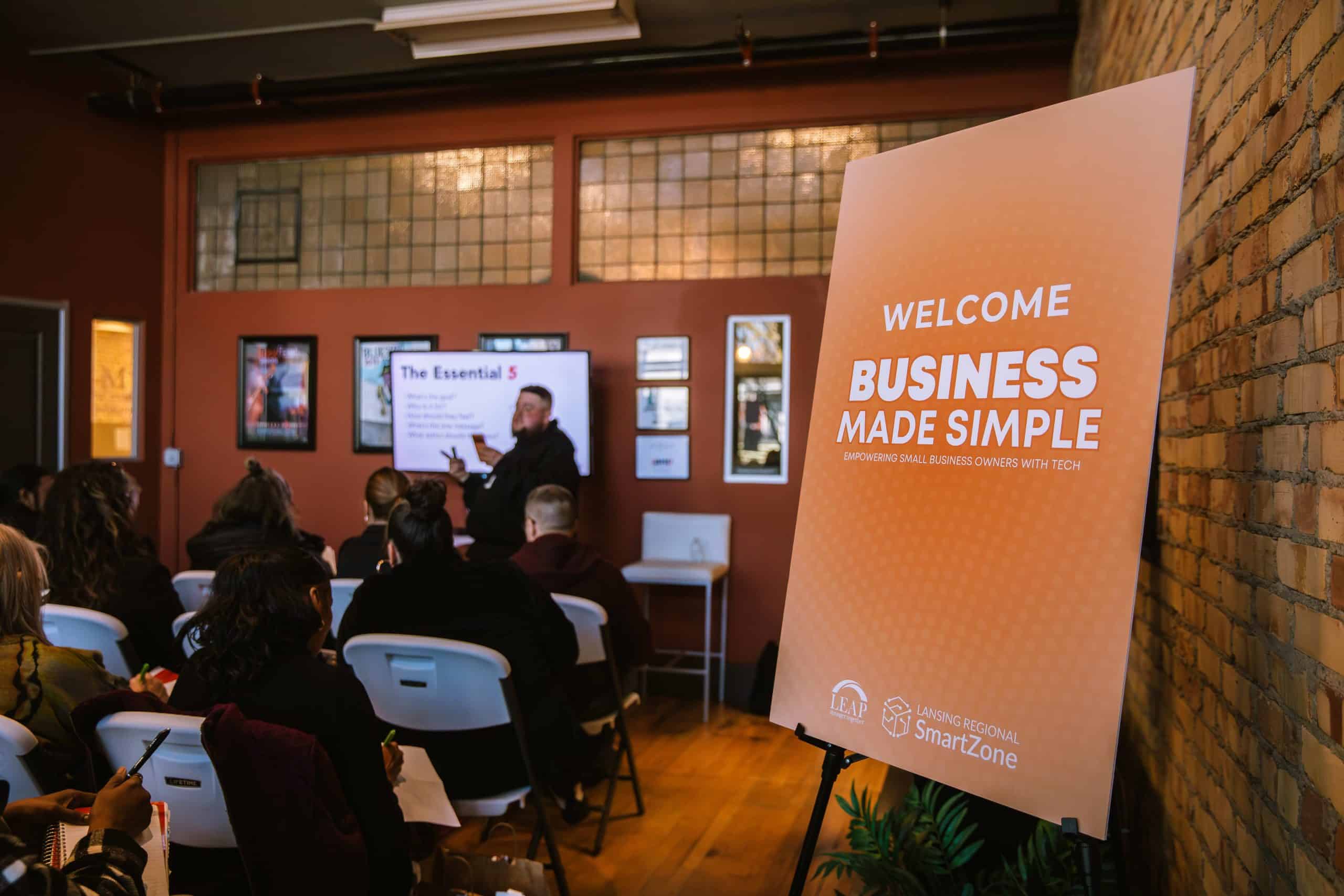A "Business Made Simple" welcome sign at a LEAP and Lansing Regional SmartZone workshop where an audience listens to a presenter.