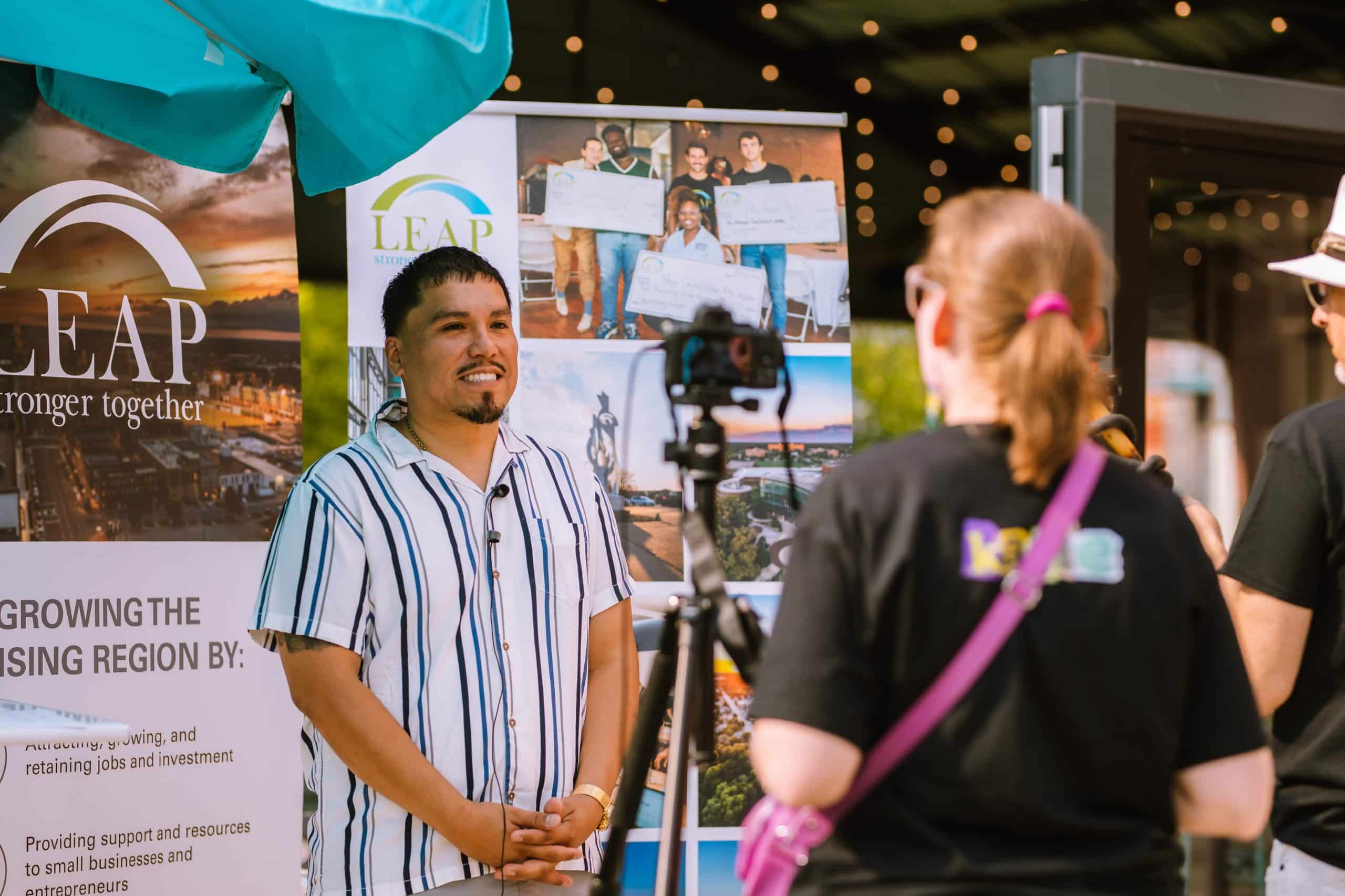 A man being interviewed on camera in front of a LEAP "Stronger Together" banner at a community event.